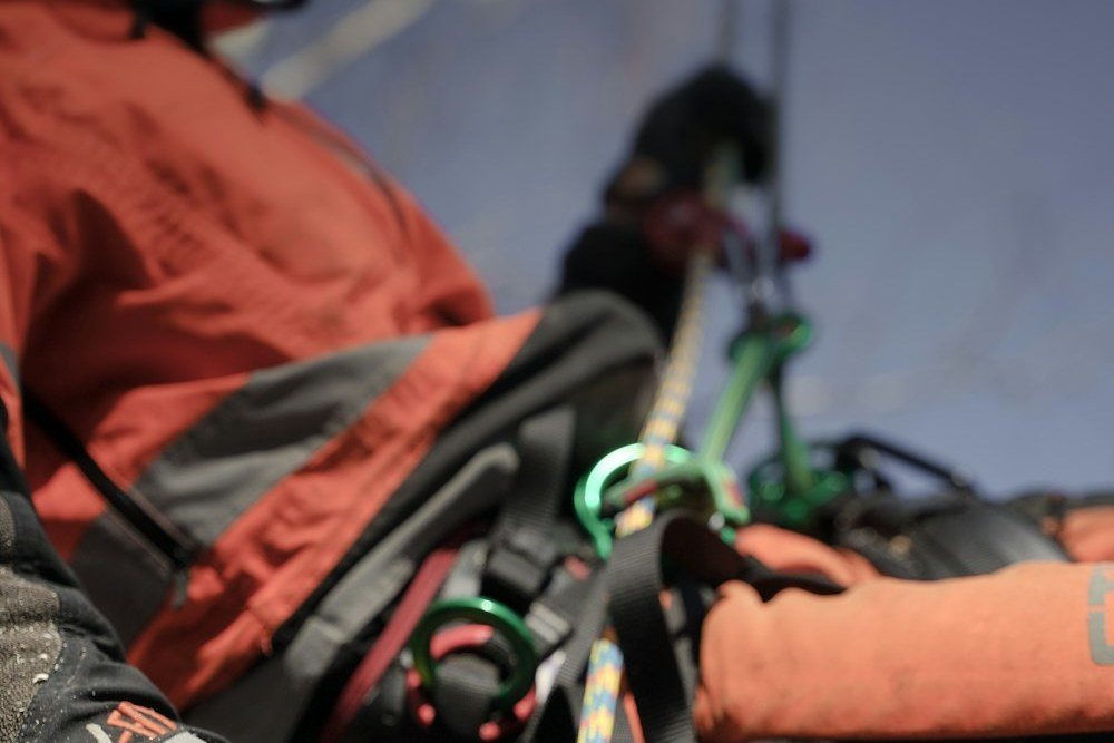 A climber in a helmet and safety glasses roped in against a trunk, reaching for a limb
