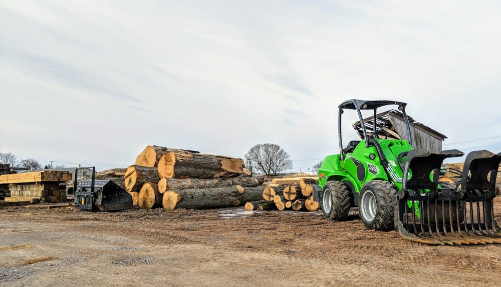 A compact track loader parked at the edge of a field beside a hardwood tree line